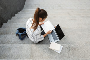 Image of person sitting on concrete steps and using a laptop and notebook. This image may depict someone who has used therapy for young adults in Philadelphia, PA for support during life transitions. A young adult therapist can support you in many ways. | 19312 | 19355